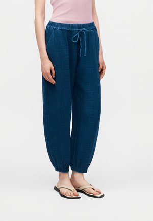 Γυναίκα που φοράει χαλαρό σκουρό μπλε υφασμάτινο παντελόνι με ελαστικά μανσέτα στους αστραγάλους και κορδόνι στη μέση, μπεζ σαγιονάρες και ροζ αμάνικη μπλούζα.