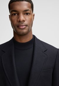 Granatowa marynarka z ostrymi klapami na czarnym, teksturowanym golfie. Widok z bliska podkreślający tkaniny i czyste linie.