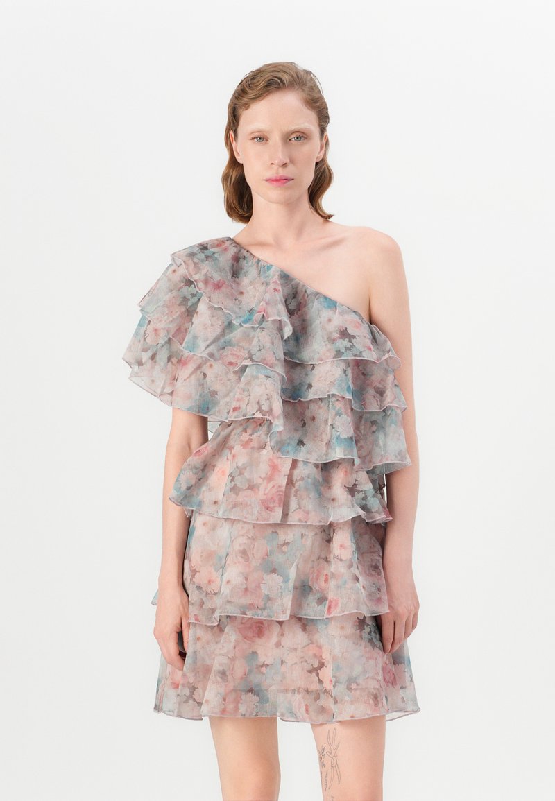 Kvinna klädd i en enaxlad, lager-på-lager blommig klänning i pastellfärger, stående mot en enkel vit bakgrund.