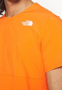 Camiseta de manga corta de color naranja brillante, hecha de una tela suave, presenta un panel texturizado en la parte inferior y un logo blanco en la parte superior izquierda.