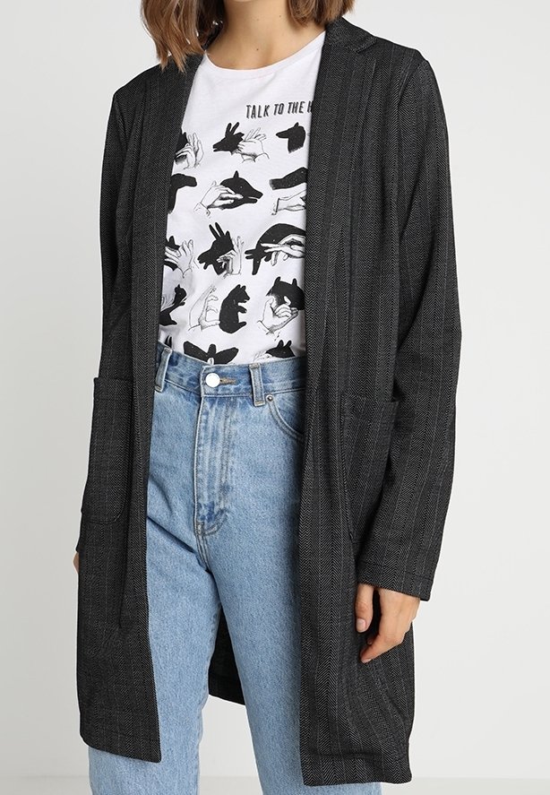Long manteau noir à motif chevrons avec devant ouvert, deux poches, porté sur un t-shirt blanc à motif graphique, associé à un jean bleu clair taille haute.