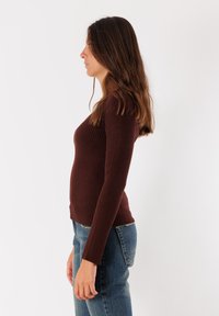 Maglietta marrone a righe con maniche lunghe e design aderente, abbinata a jeans azzurri a gamba dritta, mostrata di lato su uno sfondo bianco.