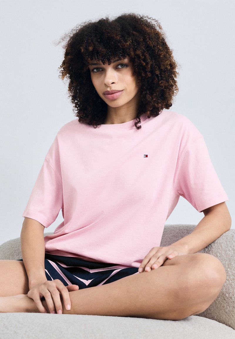 Leichtes rosa Baumwoll-T-Shirt mit kurzen Ärmeln, rundem Ausschnitt und einem kleinen Logo auf der linken Brust. Kombiniert mit gemusterten Shorts.