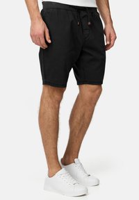 Zwarte katoenen shorts met een elastische tailleband en trekkoord, voorzien van zijzakken en zichtbare stikdetails, gedragen met witte sneakers.