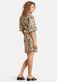 Korte mouwen jurk in een beige stof met een zwart bladpatroon, met een getailleerde taille, knielange zoom, gecombineerd met zwarte blokhakken sandaaltjes.