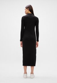 Μαύρο φόρεμα με μακριά μανίκια, κατά μήκος του γόνατου, με κολακευτική σχεδίαση, ευθεία ραφή στο πίσω μέρος και πλαϊνές σχισμές, συνδυασμένο με λευκά αθλητικά παπούτσια.