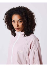 Chaqueta cortavientos de color rosa claro con cuello alto, cremallera completa y detalles en blanco. El material parece liso y de textura ligera.