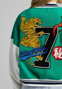 La giacca bomber verde presenta un grande ricamo di una tigre gialla, un '7' nero, il testo blu 'Stay Wild' e un emblematico cerchio rosso con simboli bianchi.