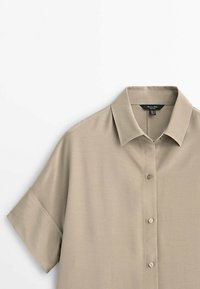 Korte mouwen, overhemd met kraag in lichtbeige stof. Heeft een knoopsluiting aan de voorkant, zachte textuur en een ontspannen pasvorm.