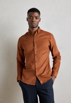 Brown button-up shirt with a collar, made of textured fabric, featuring a relaxed fit and dark buttons, worn with dark trousers.