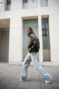 Giacca di pelle grigia oversize, jeans a vita alta blu chiaro e sandali con tacco alto bianchi. Camminando su un pavimento di pietra vicino a un edificio moderno.