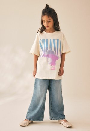 Ung pige med halvopsat hår iført overdimensioneret hvid T-shirt med teksten "SHANIA" og grafik, jeans med vide ben og glitrende beige ballerinaer.