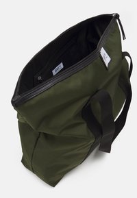 Olivgrön tygväska i slitstarkt nylon, med svarta handtag och dragkedja. Insidan innehåller en liten ficka med dragkedja och en etikett.