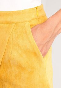 Gula mockabyxor med en slät yta, utrustade med sidofickor och en synlig dragkedja i midjan för stängning.