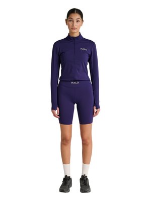 Kobieta stoi twarzą do przodu, ubrana w pasującą ciemnofioletową kurtkę zapinaną na zamek oraz dopasowane szorty, białe skarpetki i czarne buty sportowe.