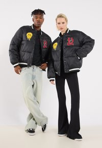 Black puffer jackets with embroidered designs, including a yellow smiley face and red lettering. Standalone with ribbed cuffs and hem.