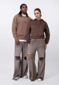 Pruun fliiskampsun, lühendatud pruuni kapuutsiga dressipluus köieotstega ja beežid laiade säärtega püksid rebitud detailidega; vabaajaspordiriietus, neutraalsed toonid.