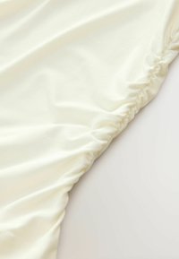 Kremo spalvos audinys su minkšta tekstūra, su susiraukšlėjimo detalėmis palei vieną kraštą, sukuriantis lygų, krentančio elegancijos efektą.
