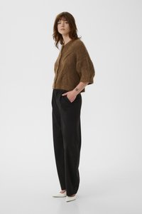 Γυναίκα με καφέ μάλλινη ζακέτα, μαύρο παντελόνι φαρδύ, λευκά μυτερά τακούνια, στέκεται με το ένα χέρι στην τσέπη μπροστά σε απλό φόντο.