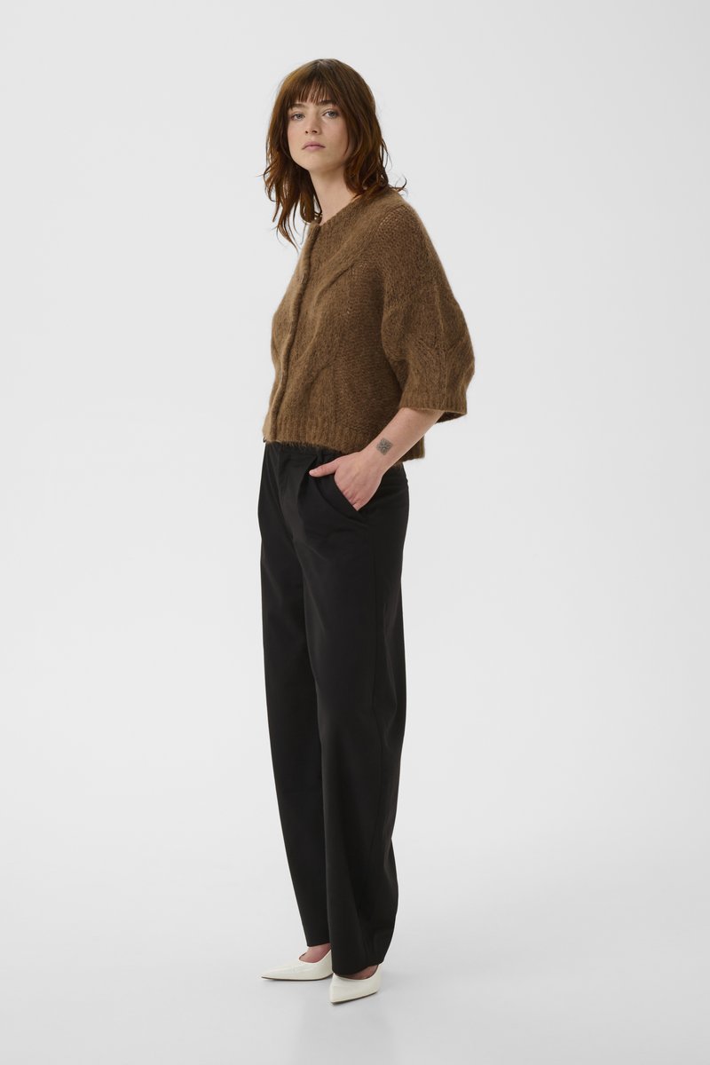 Femme portant un cardigan marron pelucheux, un pantalon noir à jambes larges, des talons blancs pointus, debout avec une main dans la poche devant un fond uni.