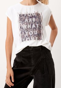 Žena v bílém tričku s nápisem "To, co miluješ, tě definuje", podloženým vzorovaným čtvercem, ve spojením s černými kalhotami s vysokým pasem.