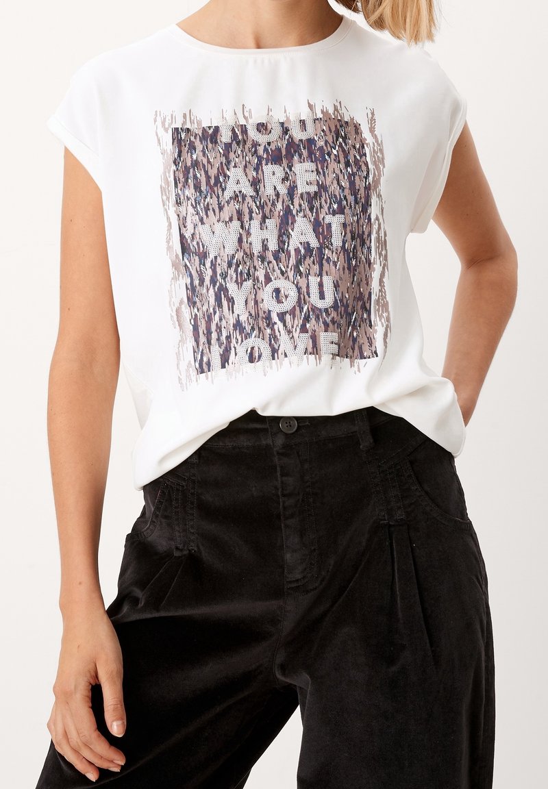 Žena v bílém tričku s nápisem "To, co miluješ, tě definuje", podloženým vzorovaným čtvercem, ve spojením s černými kalhotami s vysokým pasem.