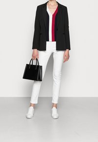 Frau in einem schwarzen Blazer, gestreiftem V-Ausschnitt-Oberteil, weißen Hosen und weißen Sneakers, die eine schwarze Handtasche vor einem weißen Hintergrund hält.