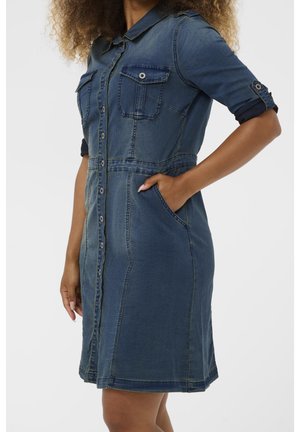 Kvinde iført en blå denim knap-foret kjole med opkørte ærmer og lommer, stående med den ene hånd i en lomme.
