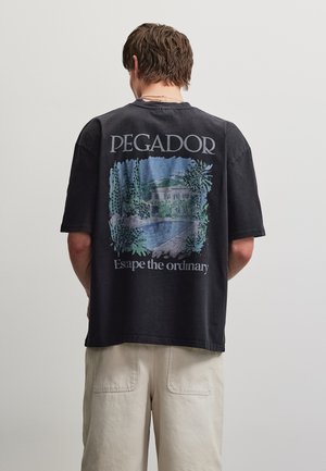 Homme aux cheveux bruns courts portant un T-shirt noir avec un graphisme de maison tropicale et piscine et le texte « PEGADOR Escape the ordinary. »
