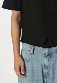 Gilet in lana verde scuro con orlo arrotondato, caratterizzato da una parte anteriore divisa, sei bottoni e due tasche laterali, abbinato a jeans denim blu sbiaditi.