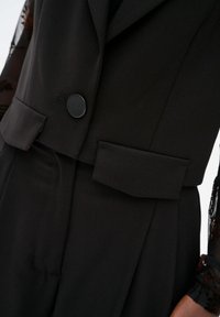 Czarna dopasowana marynarka z pojedynczym okrągłym guzikiem, wyraźnie zarysowanymi ramionami i podwójnymi klapkami kieszeni. Gładki materiał z lekkim połyskiem.