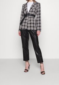 Blazer a cuadros en blanco y negro con una cintura entallada y cinturón integrado, combinado con pantalones de cuero sintético negros y tacones negros con tiras.