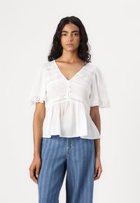 Bílá peplum halenka s krátkými nafouknutými rukávy; má krajkový lem a zapínání na knoflíky; je kombinována s modrými pruhovanými širokými kalhotami.