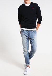 Man som bär en svart tröja över en vit skjorta, ljusblå uppvikta jeans och vita sneakers, står mot en vit bakgrund.