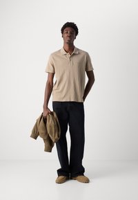Polo shirt beige in cotone con maniche corte, pantaloni a gamba larga blu scuro e un felpa marrone tenuta in una mano. Calzature beige completano il look.