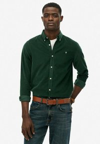 Joven vestido con una camisa de botones verde oscuro sobre una camiseta blanca, con jeans azules y un cinturón de cuero marrón, de pie con una mano detrás de la espalda.