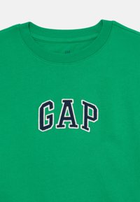 T-shirt verde de algodão com decote redondo e logótipo "GAP" bordado em azul-marinho e branco. Textura suave com um acabamento em cor sólida.
