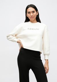 Sweat-shirt blanc avec "ARMANI" en lettres argentées, doté d'un col rond et de manches bouffantes, assorti à un pantalon noir côtelé.