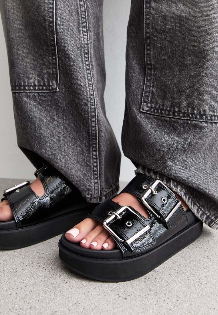 Sandali platform neri con due cinturini con fibbie, caratterizzati da una finitura lucida e una suola spessa e solida nera. Abbinati a pantaloni larghi grigi.