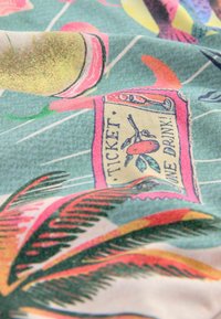 Tkanina z kolorowym tropikalnym wzorem, na którym znajdują się liście palm, napoje oraz wzór biletu. Dominującymi kolorami są zieleń, róż i żółć.