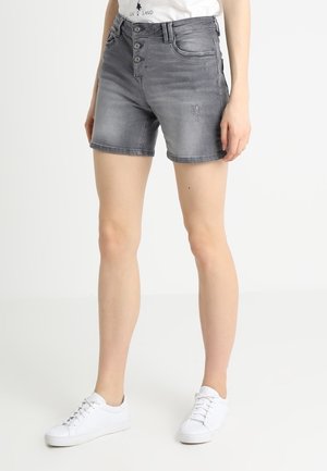Kvinde ben iført grå denim shorts med knaplukning, hvide sneakers og en hvid skjorte lagt ned i bukserne, stående mod en ensfarvet baggrund.