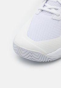 Vit atletikssko med en texturerad mesh-övre, släta syntetiska detaljer och en gummisula med ett zigzag-mönster.