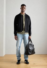 Black bomber jacket, olive green shirt, light blue jeans, and black sneakers. Holding a black leather bag with a zipper and handles.