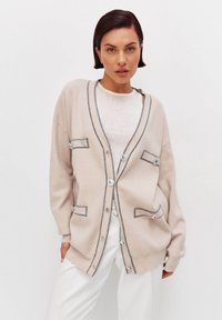 Cárdigan beige con costuras grises, de corte oversize, con grandes bolsillos frontales y acentos de botones plateados. Llevado sobre una blusa blanca.