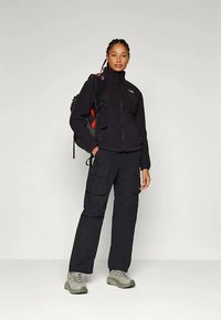 Naine musta jope ja cargo-pükstega, seljas matkasussid ja kandes musta-punast seljakotti, seisab ühtlasel taustal.