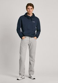 Jūras zila kapuce ar aukliņu un priekšējo kabatu, saskaņota ar gaiši pelēkiem džinsiem un baltiem sporta apaviem ar pelēkiem akcentiem.