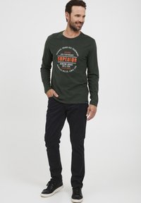 Grøn skjorte med lange ærmer og en cirkulær grafik i orange og hvid. Båret med mørke bukser og sorte sneakers, hvilket giver et afslappet look.