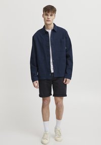 Navy blue, button-up jacket with a single chest pocket, made of cotton, paired with black shorts and white socks, worn with sneakers.