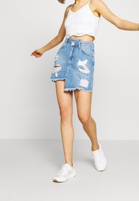 Denim-Minirock mit einem verwaschenen Design, fransigem Saum, mehreren Rissen und hoher Taille. Kombiniert mit einem gekürzten weißen Spitzentop und Sneakers.