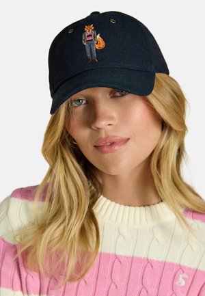 Donna bionda che indossa un berretto nero con una volpe ricamata, in abbigliamento casual, incluso un maglione a righe bianco e rosa.